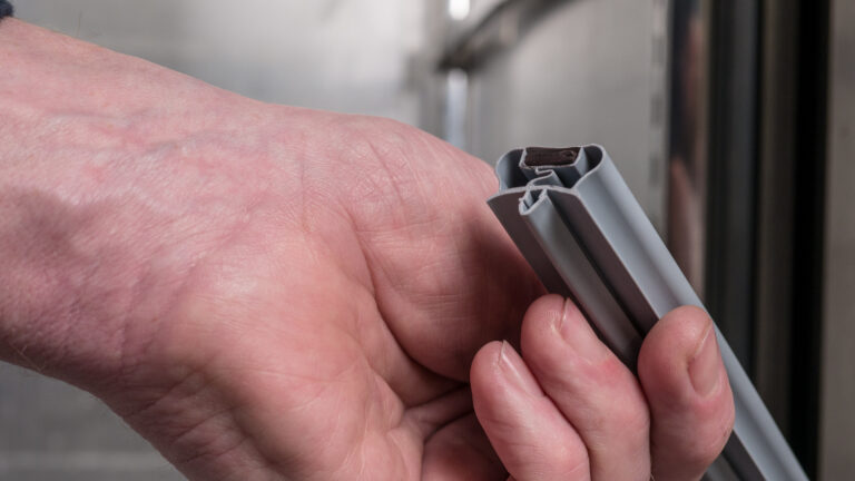 A close-up of a person's hand holding a grey rubber or plastic sealing strip, likely used for insulation or weatherproofing around doors or windows. The background is blurred, focusing attention on the hand and the strip.