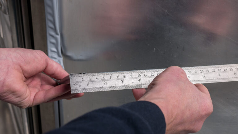 A close-up of two hands holding a metal ruler against a shiny, metallic or glass surface. The ruler is marked in both inches and centimetres, and the person appears to be measuring the edge of the surface. The background shows reflections and a taped seam.