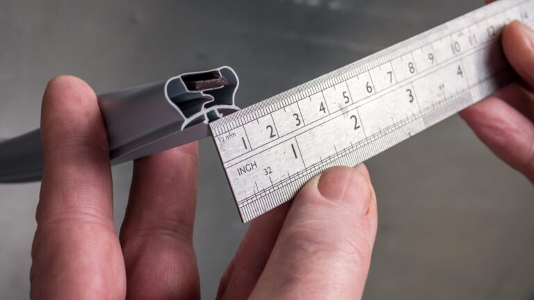 A close-up of hands holding a metal ruler to measure the width of a grey rubber or plastic seal with a hollow profile, against a neutral background. The ruler markings and the detailed profile of the seal are clearly visible.