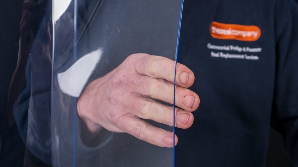 A close-up of a person’s hand holding a clear acrylic sheet. The person is wearing a dark blue shirt with an orange and white logo on the left chest, partially reading “Glassalcompany” and mentioning commercial glazing and replacement services.