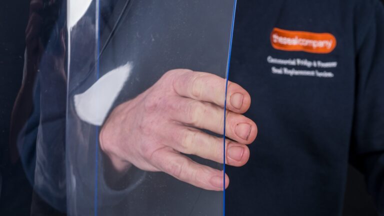 A close-up of a person’s hand holding a clear acrylic sheet. The person is wearing a dark blue shirt with an orange and white logo on the left chest, partially reading “Glassalcompany” and mentioning commercial glazing and replacement services.