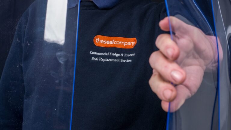 A person in a navy shirt with an orange “thesealcompany” logo and text reading “Commercial Fridge & Freezer Seal Replacement Services” pulls a clear plastic strip, partially obscuring their face with their hand and the strip.