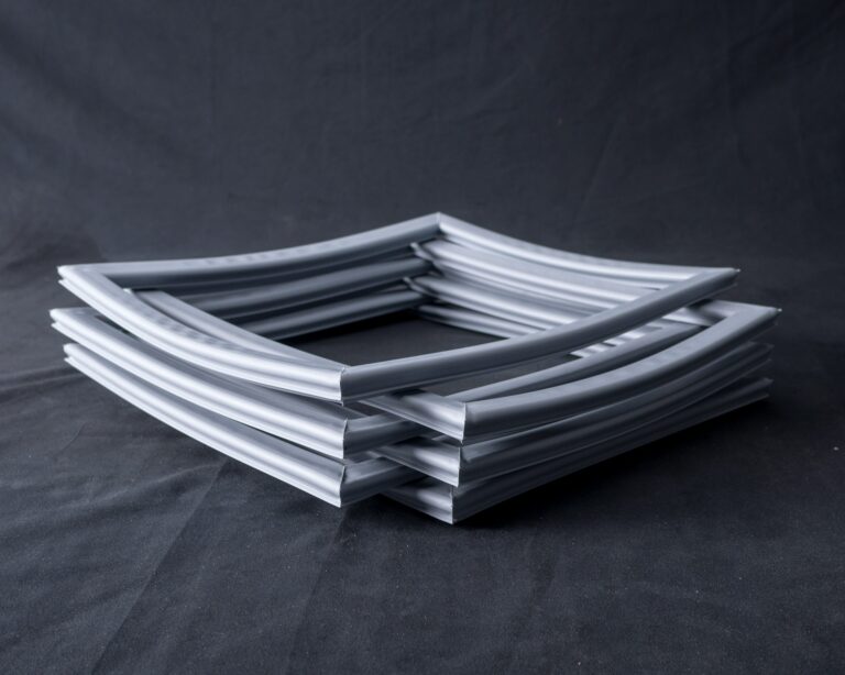 A stack of four square, grey, flexible rubber gaskets arranged neatly on a dark fabric background. The gaskets have a groove along their length and are stacked in an alternating, offset pattern.