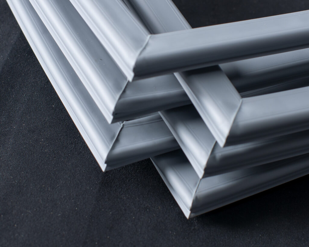 Three grey picture frames intersect and overlap at varying angles on a black surface, forming a geometric arrangement with their corners. The frames create a layered, abstract pattern with smooth, matte finishes and clean lines.