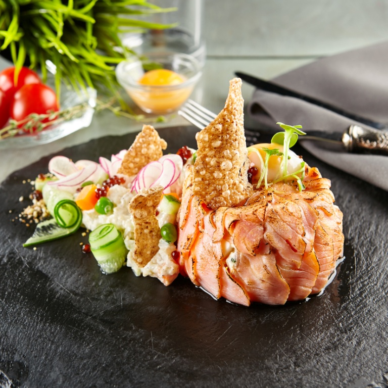A gourmet dish of thinly sliced, wrapped salmon garnished with microgreens, crispy crackers, cucumber ribbons, radish slices, peas, and a creamy salad, artfully plated on a round slate, with a grey serviette and decorative ingredients in the background.