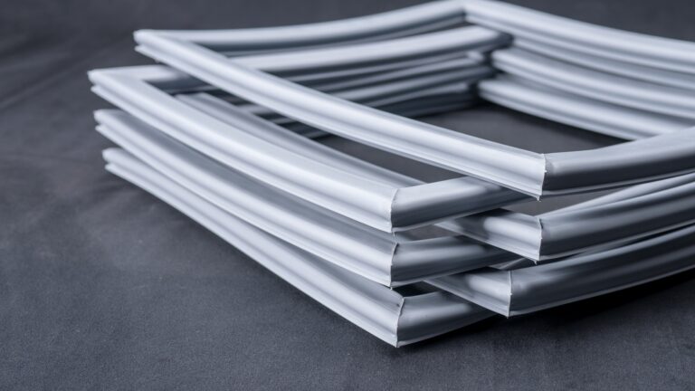Four rectangular, grey rubber or plastic gaskets are stacked in an alternating criss-cross pattern on a dark grey surface. The gaskets have smooth surfaces and rounded edges, forming an orderly, geometric arrangement.