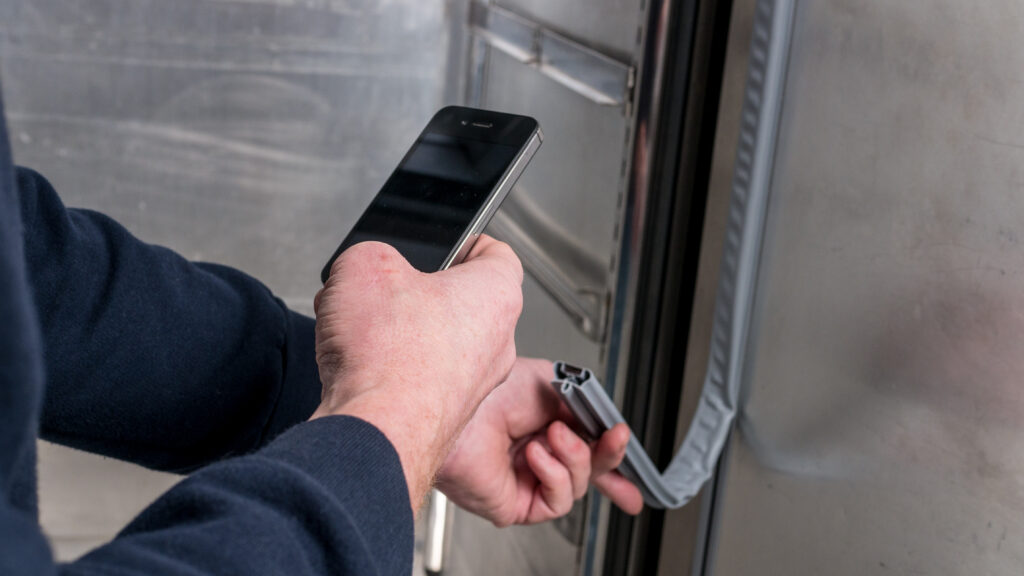 A person wearing a dark long-sleeve shirt holds a mobile in one hand and inspects the rubber seal on the edge of a metallic door with the other hand. The setting appears industrial or commercial, with stainless steel surfaces visible.