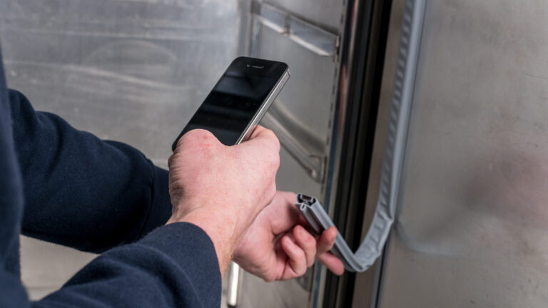 A person wearing a dark long-sleeve shirt holds a mobile in one hand and inspects the rubber seal on the edge of a metallic door with the other hand. The setting appears industrial or commercial, with stainless steel surfaces visible.