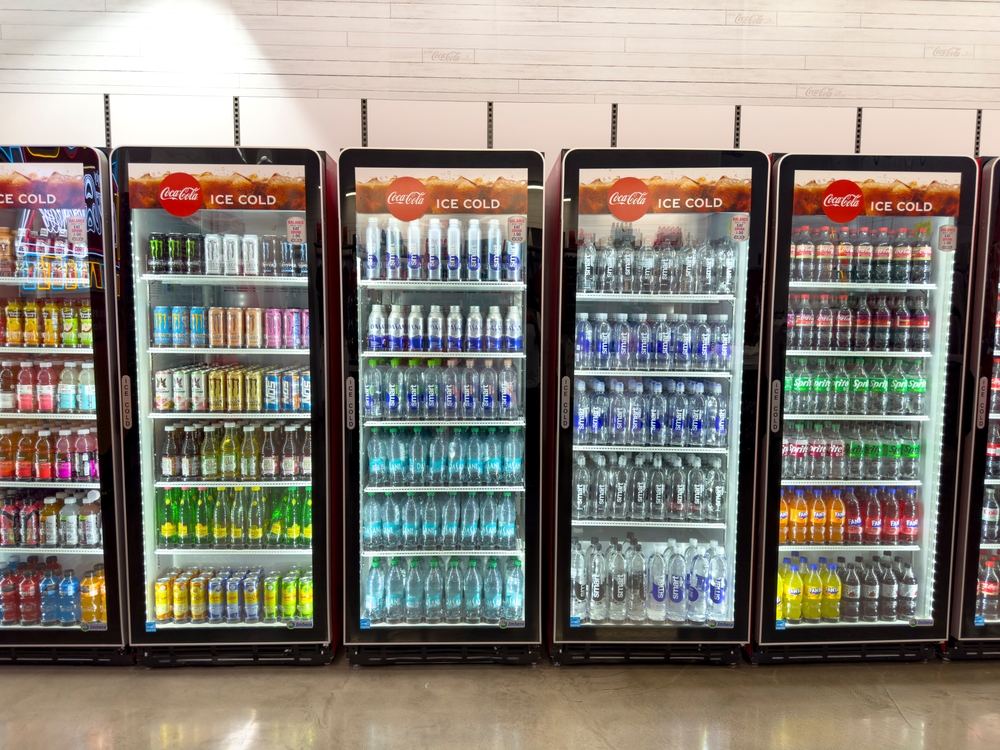 Four glass-door fridges filled with various cold drinks, including fizzy drinks, bottled water, sports drinks, and juices, are lined up in a shop. The fridges are labelled Coca-Cola ICE COLD with bright lighting highlighting the colourful bottles and cans.