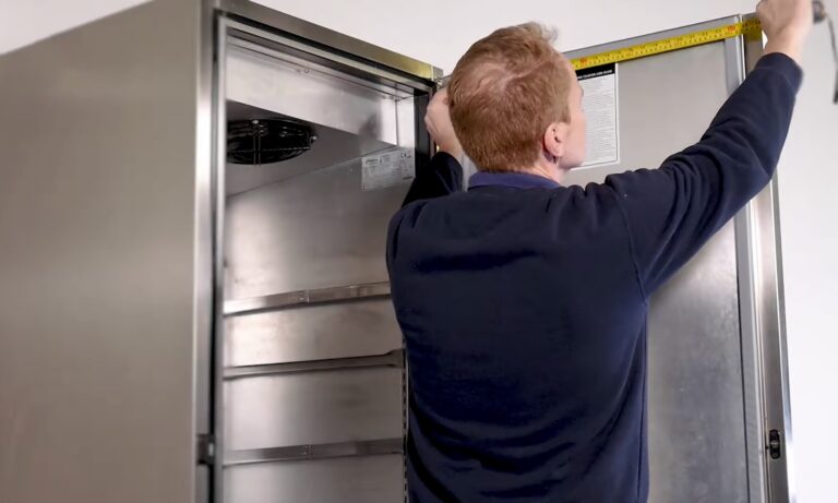 A man in a navy jumper uses a yellow tape measure to measure the height of an open, empty stainless steel fridge, reaching from the top edge to the bottom inside corner. The fridge door is fully open, showing shelves inside.