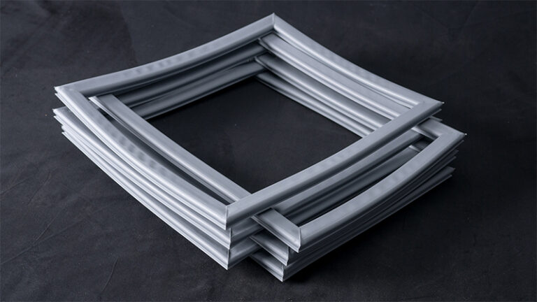 Four square, flexible, grey rubber or silicone gaskets are stacked and slightly askew on a dark surface. Each gasket has a ridged texture and the arrangement creates a layered, geometric pattern with shadowed edges.