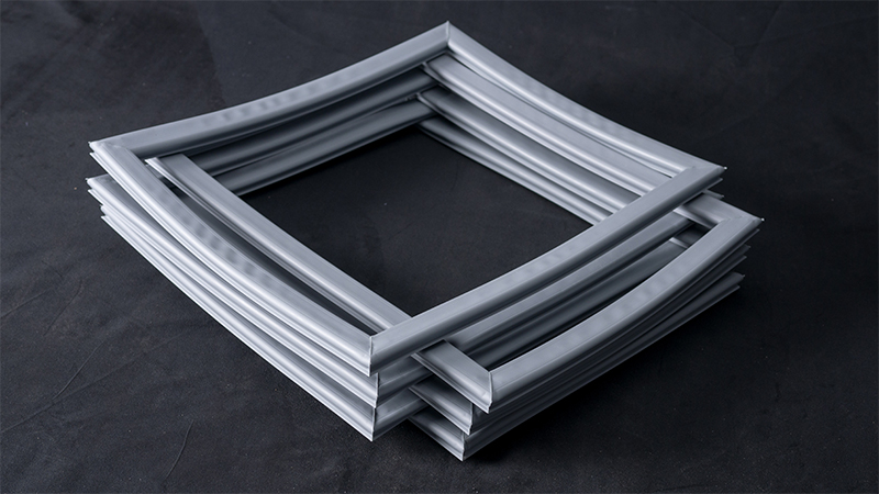 Four square, flexible, grey rubber or silicone gaskets are stacked and slightly askew on a dark surface. Each gasket has a ridged texture and the arrangement creates a layered, geometric pattern with shadowed edges.
