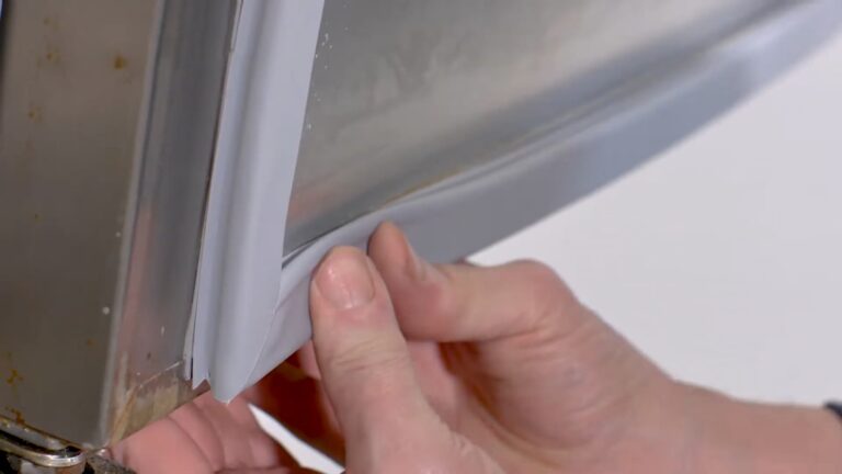 Close-up of a person's hands installing a flexible, light grey rubber seal onto the edge of a metal appliance door, possibly a refrigerator or freezer, with the white background out of focus. The person carefully lines up the gasket along the door edge.