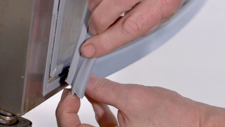 A close-up of two hands installing a flexible grey rubber gasket onto the edge of a metal appliance door, possibly a refrigerator or freezer. The hands press the gasket firmly into the groove, ensuring a tight seal. The background is out of focus and light-coloured.