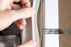 A close-up of a person's hand pulling back the rubber seal on a fridge door, inspecting the gasket for cleanliness or damage. The fridge interior and a transparent container are partially visible. The focus is on the seal and the fingertips.