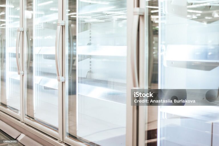 A row of glass-door fridge units in a brightly lit supermarket, all shelves inside are empty. The scene appears clean and sterile, with reflections on the glass doors. No products or people are visible.