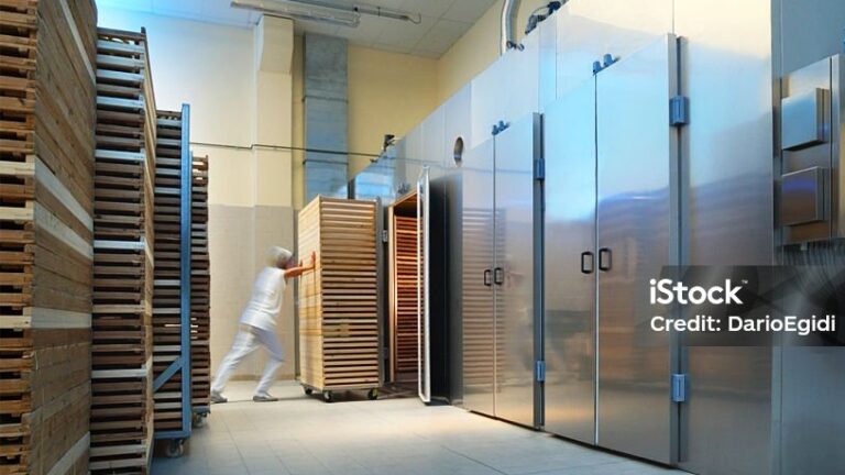 A worker in white clothing pushes a tall rack of wooden trays into a large, stainless steel industrial oven or drying chamber in a clean, well-lit food processing facility. Rows of similar racks are lined up on the left. Image credit: DarioEgidi/iStock.