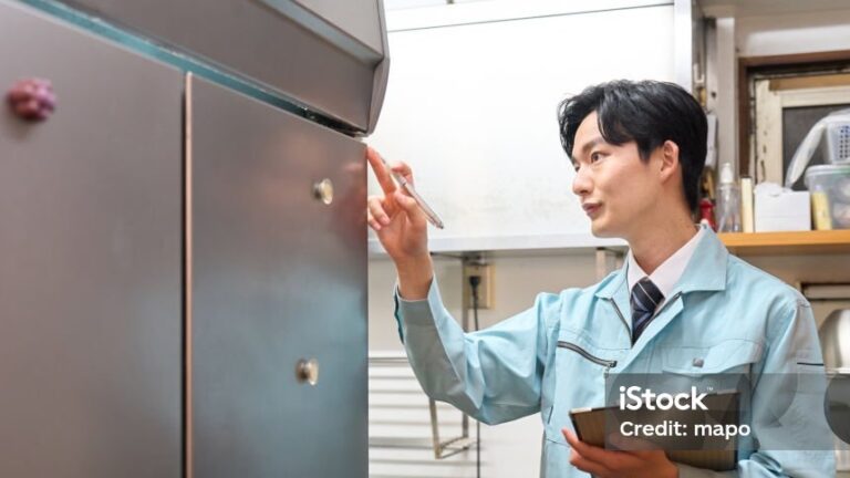 A man in a light blue uniform holds a tablet whilst inspecting a metallic fridge in a kitchen. He points at the fridge’s door, focused on his task. Shelves with crockery and containers are visible in the background.
