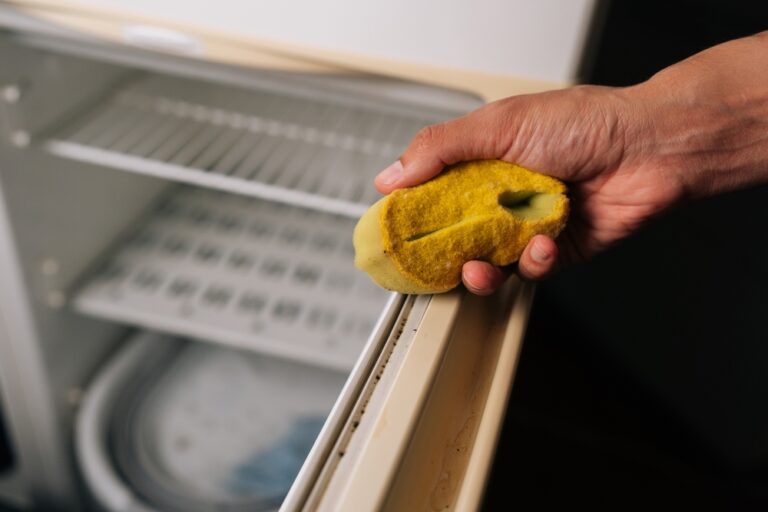 A hand holds a yellow sponge, cleaning the rubber seal on the open door of an empty fridge. The fridge interior is visible, showing empty shelves and a clean, white surface. The focus is on the cleaning action at the fridge door seal.