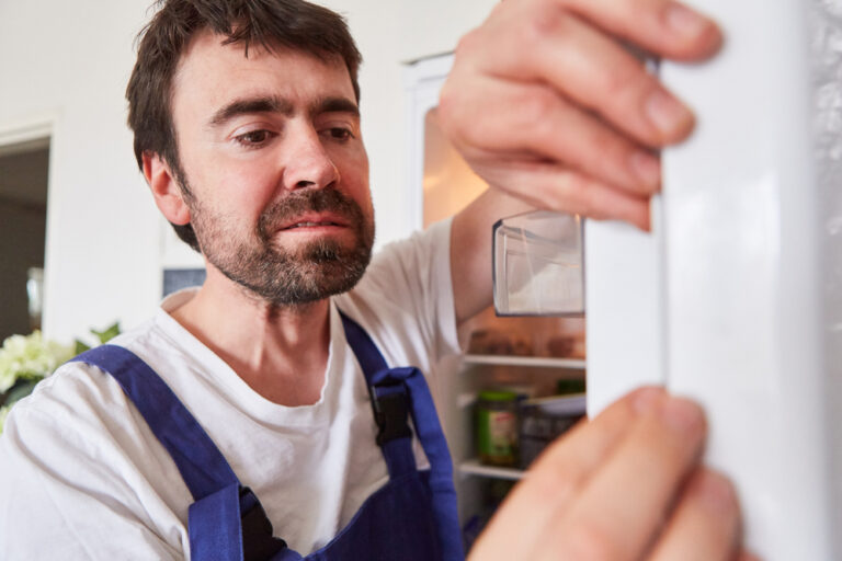 A man in blue overalls and a white shirt repairs or inspects the seal of an open fridge door, focusing intently on his task. The fridge interior with food and containers is partially visible in the background.