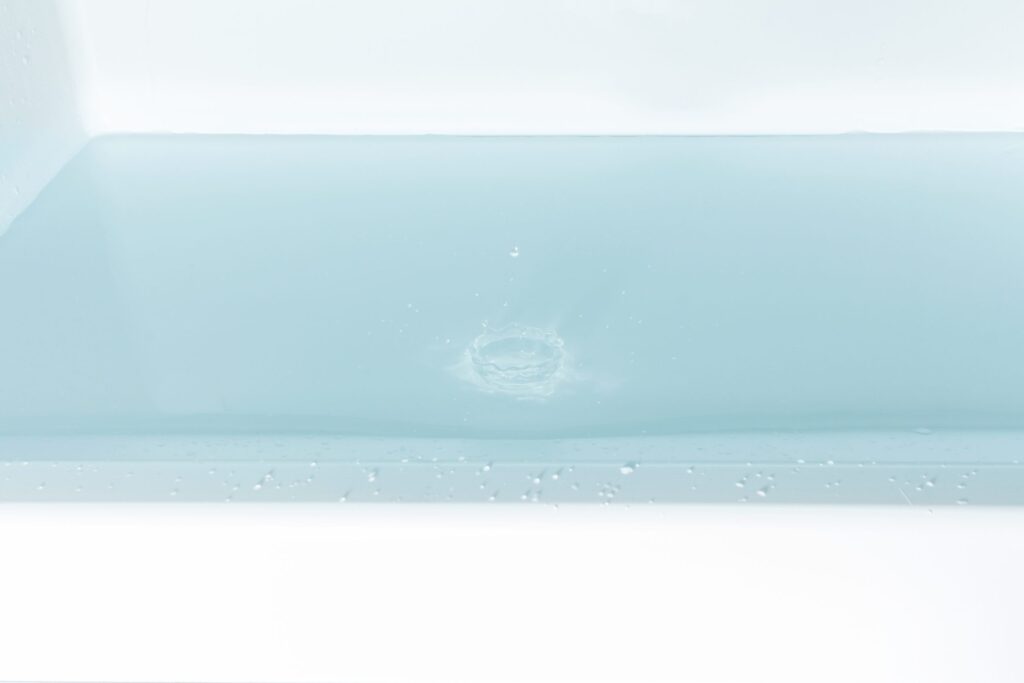 A close-up of a bath filled with clear, pale blue water. A small splash and ripples are visible at the water’s surface, suggesting something has just dropped in. The bath’s clean, white edge is visible at the bottom of the image.