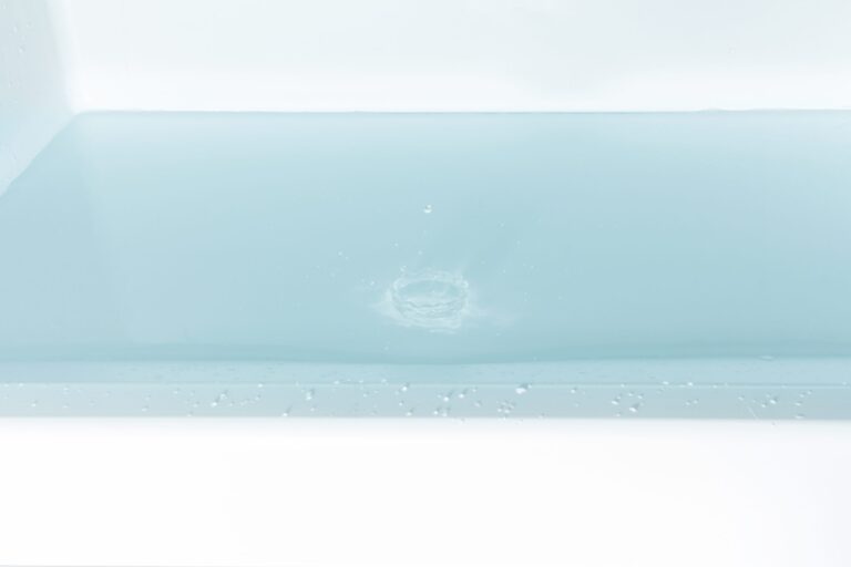 A close-up of a bath filled with clear, pale blue water. A small splash and ripples are visible at the water’s surface, suggesting something has just dropped in. The bath’s clean, white edge is visible at the bottom of the image.
