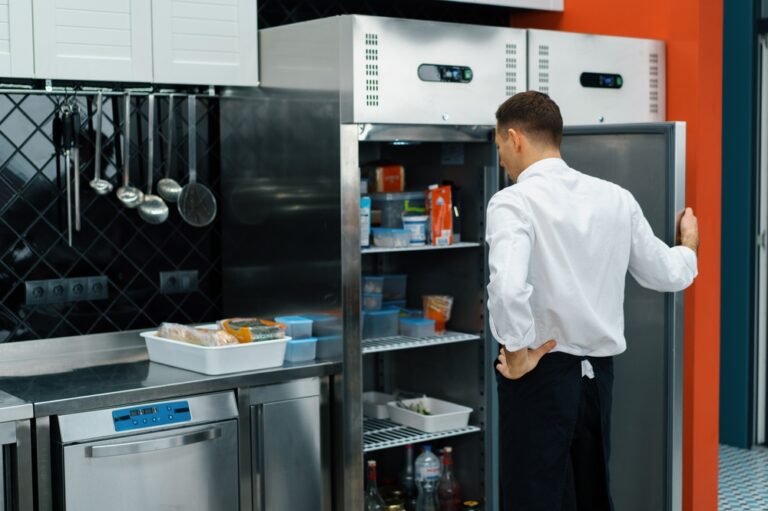 A man in a white chef’s coat stands with one hand on his hip, looking into an open commercial fridge in a modern kitchen. Shelves inside the fridge hold various food items. Utensils hang on a black tiled wall nearby.