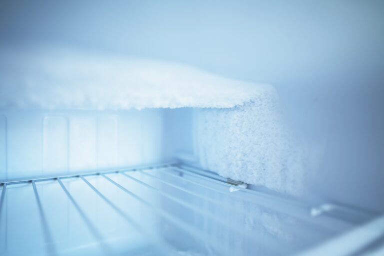The inside of a freezer with a thick layer of white frost and ice build-up on the top and back wall, above empty metal shelves. The scene is lit with a cool, bluish light, emphasising the cold, icy environment.