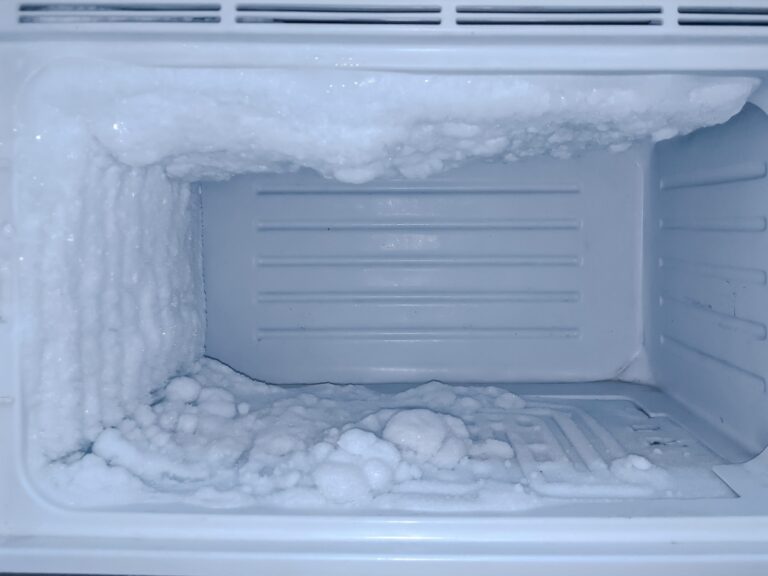 An empty freezer with heavy frost and ice build-up on the back, left wall, and floor. The thick layer of white ice appears uneven and jagged, coating much of the interior surfaces, indicating the freezer needs defrosting.