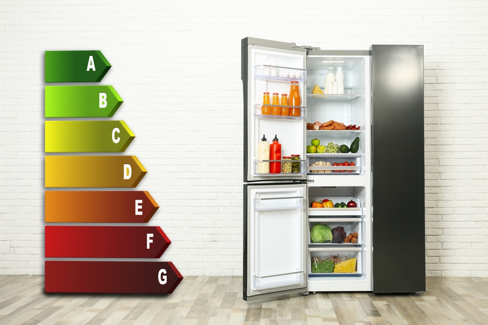 A modern fridge with its doors open, revealing organised shelves of food and drinks, stands on a wooden floor beside an energy efficiency rating chart ranging from green A (most efficient) to red G (least efficient) against a white brick wall.