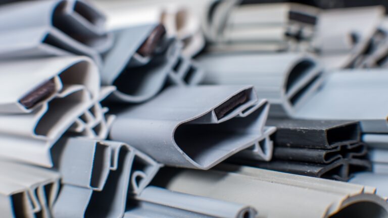 Close-up of stacked, grey and black rubber or plastic extrusions with various hollow, angular, and curved shapes. The image highlights textured surfaces and overlapping profiles, suggesting industrial or construction materials, possibly for sealing or insulation purposes.