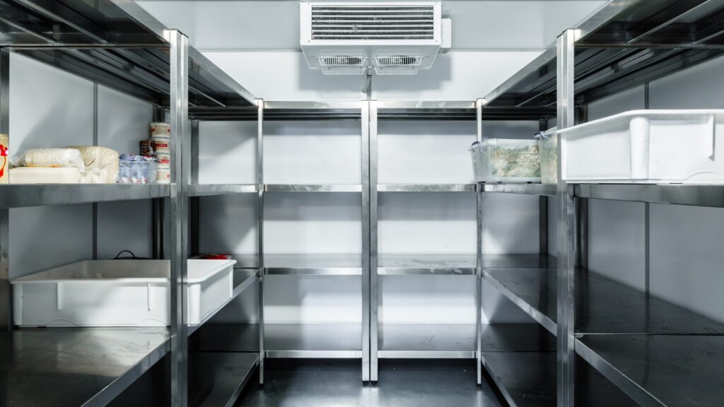 A clean, empty commercial walk-in fridge with stainless steel shelves along both sides. A few plastic bins and containers with food items are sparsely placed on the shelves, while an air vent helps minimise cold room condensation on the back wall.