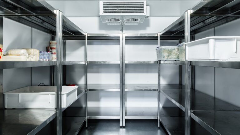 A clean, empty commercial walk-in fridge with stainless steel shelves along both sides. A few plastic bins and containers with food items are sparsely placed on the shelves, while an air vent helps minimise cold room condensation on the back wall.