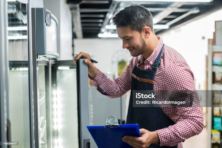 A man wearing a checked shirt and apron smiles whilst checking items inside a commercial fridge. He holds a pen and blue clipboard, possibly taking stock, in a brightly lit shop or supermarket aisle.