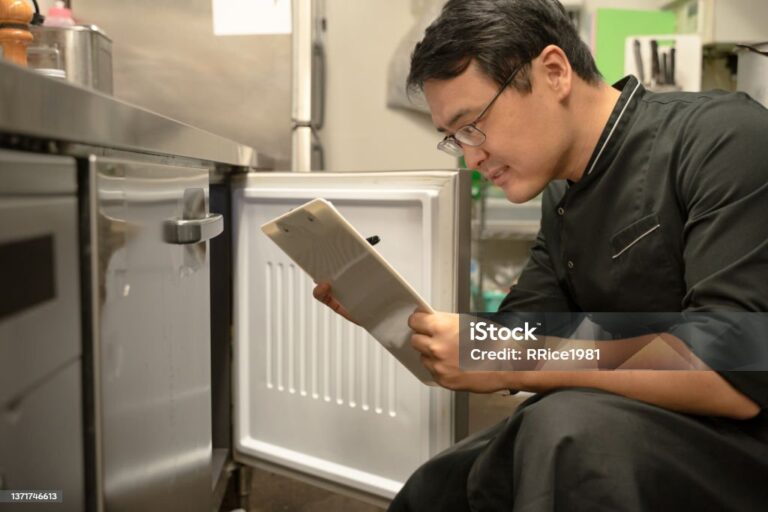 A man in glasses and a black shirt kneels in front of an open freezer, writing on a clipboard. He appears to be inspecting or taking inventory in a kitchen setting, with stainless steel appliances and shelves visible in the background.