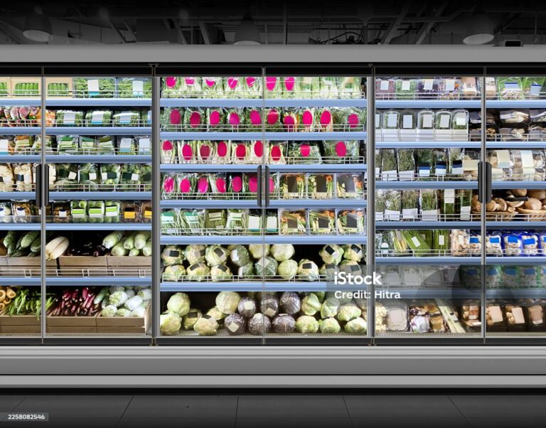 A supermarket refrigerated section with glass doors displays neatly arranged fresh vegetables, including leafy greens, cabbages, aubergines, mushrooms, packaged herbs, and dragon fruit on shelves under bright lighting.