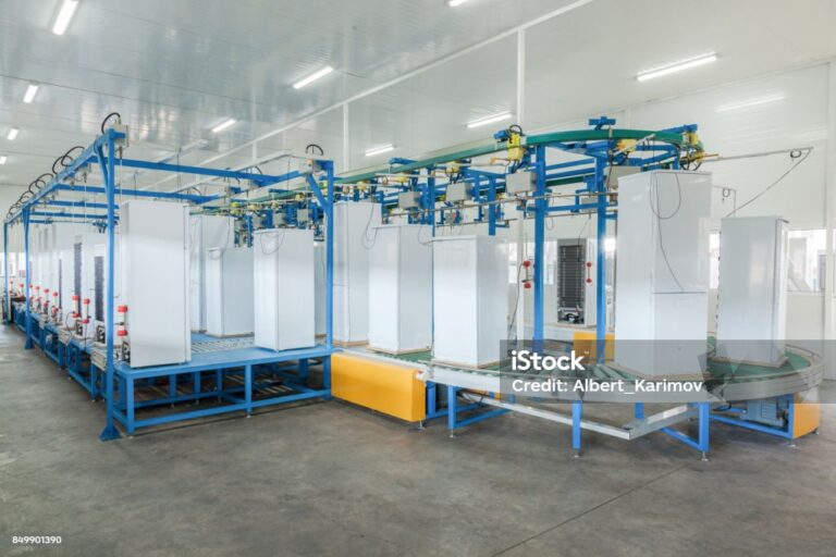 A large industrial factory interior with a blue metal conveyor system carrying white refrigerator doors. The space has bright overhead lighting, white walls, and a concrete floor. The environment is clean and organised, with no workers present.